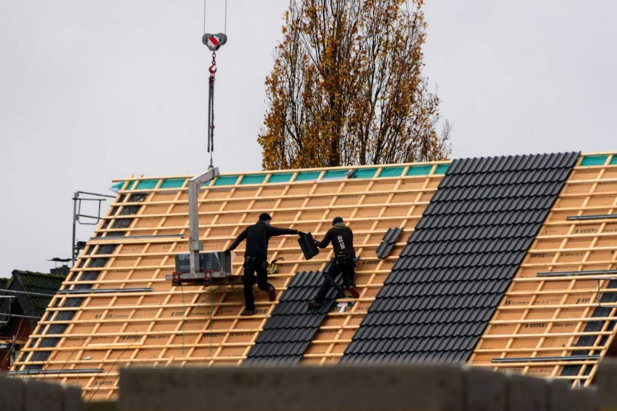 Zwei Dachdecker verlegen dunkle Dachziegel, Material hängt am Kran